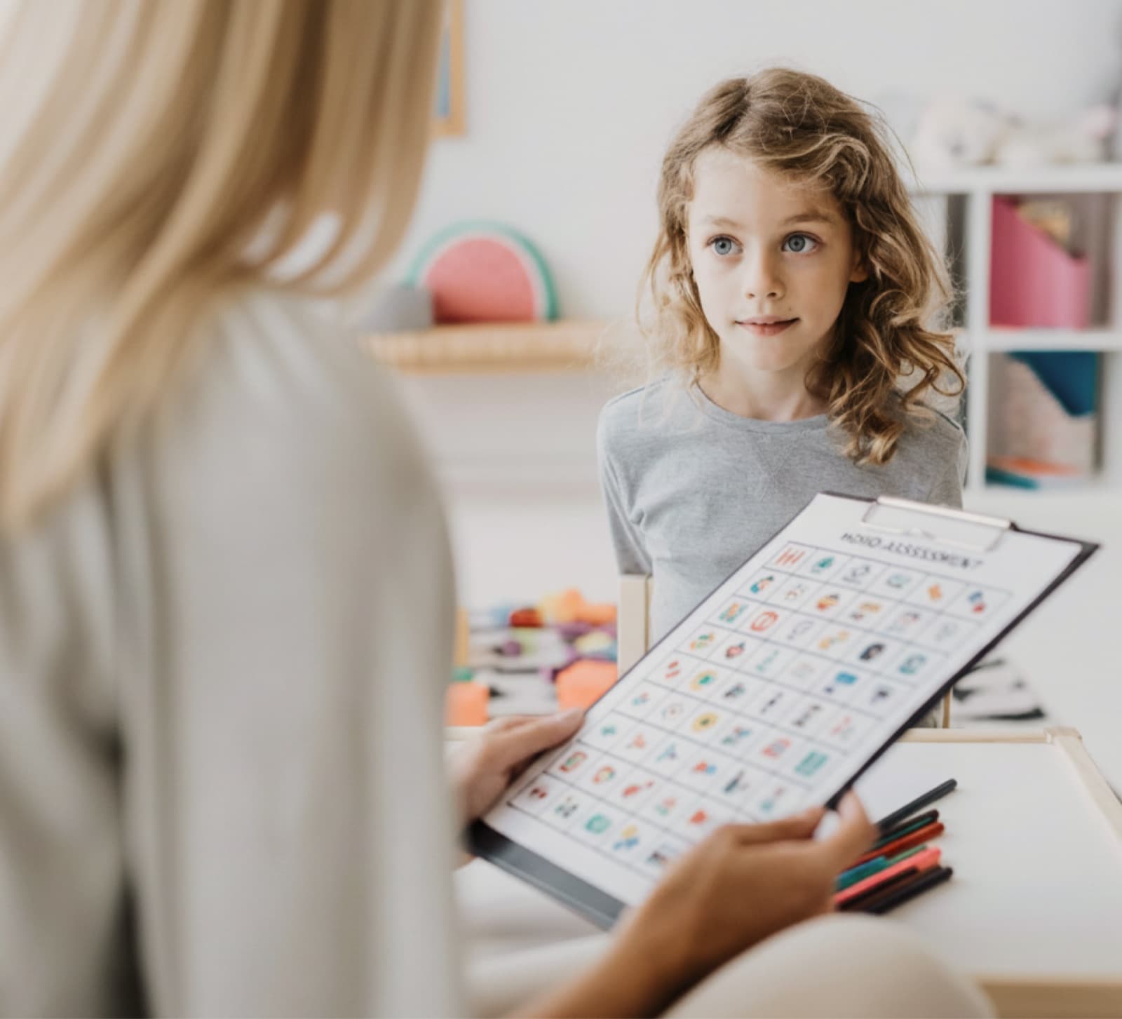 A school-age child sits across from a clinician who is holding a colourful picture-based mood-assessment chart during a session